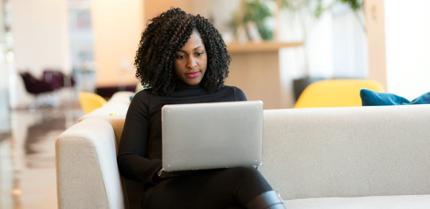 A professional woman works on a laptop while sitting comfortably on a couch in a modern office or common area.