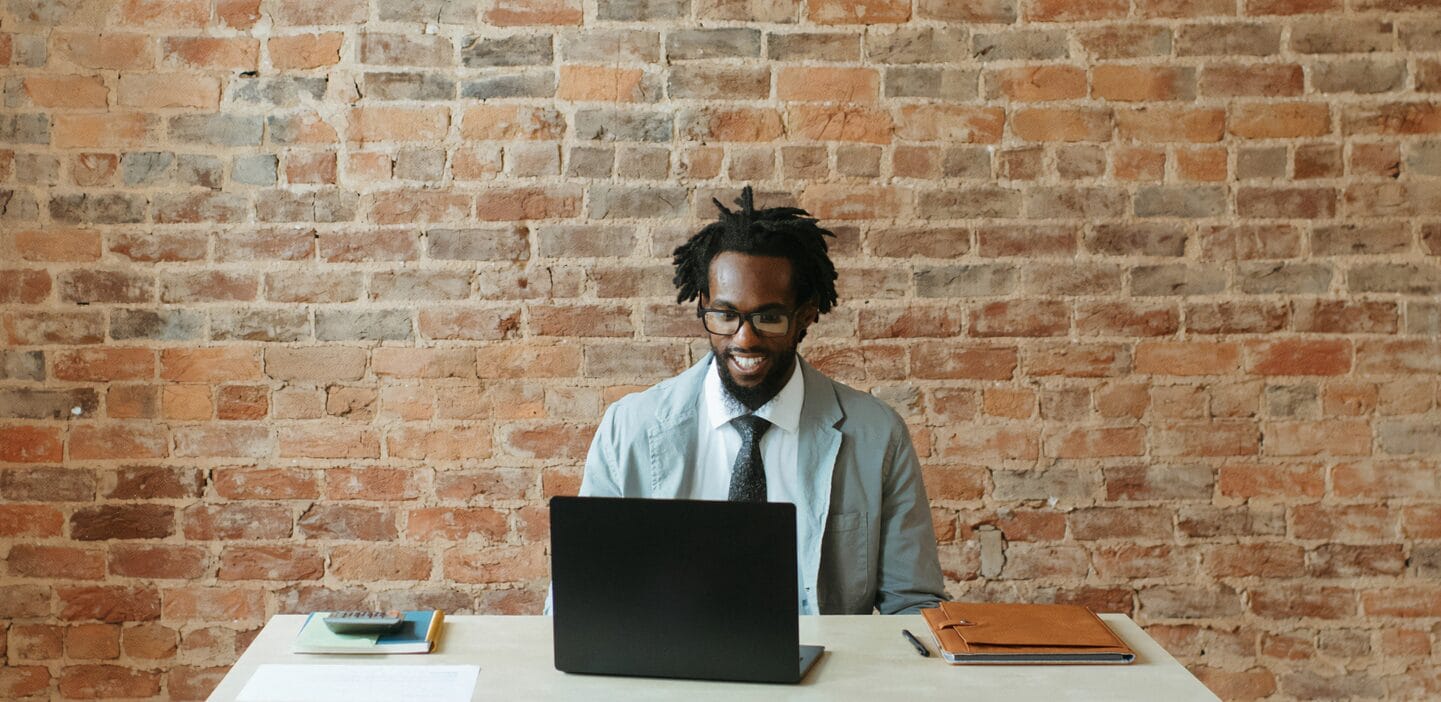 Smiling man in a gray blazer and glasses looks down at an open black laptop at a white desk. The background is a prominent red brick wall.